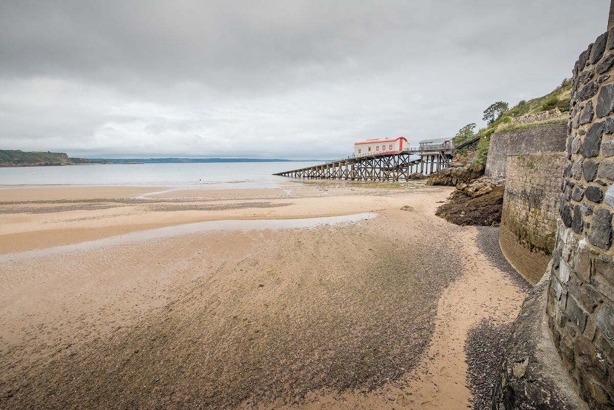 A coastal scene with a stone wall, a wooden pier, and a building with a red roof.