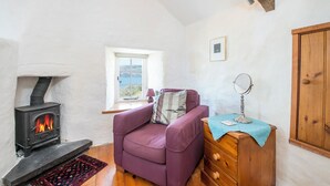 Une pièce chaleureuse avec un poêle à bois, un fauteuil violet, une commode en bois et une fenêtre donnant sur la mer.