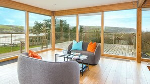 Een ruim balkon met een houten vloer, een grijze bank met oranje en blauwe kussens, en een glazen tafel met een theepot en boeken.