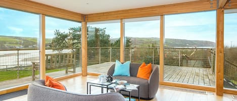A spacious balcony with a wooden floor, a grey sofa with orange and blue cushions, and a glass table with a teapot and books.
