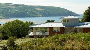 Een modern huis met grote ramen, een terras en uitzicht op strand en heuvels.