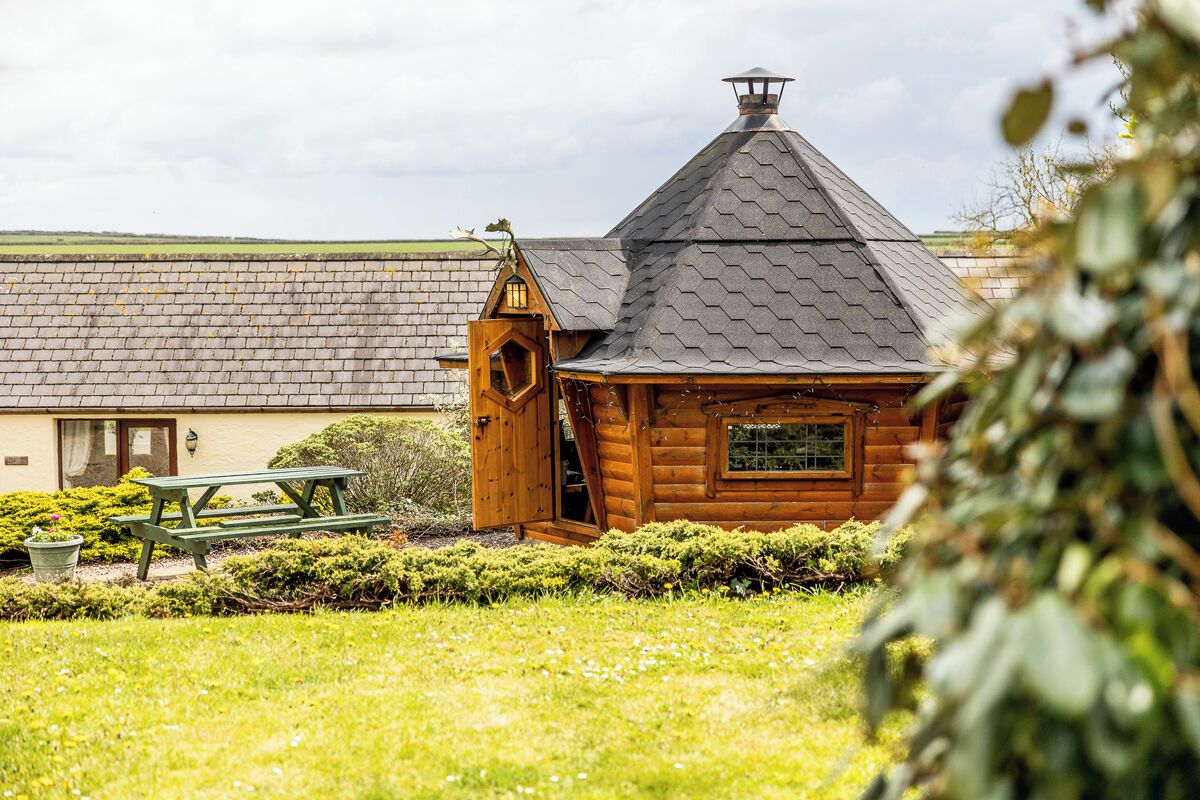 Une cabane en bois avec un toit en pente et une cheminée, entourée d’une pelouse bien entretenue et d’une table de pique-nique.