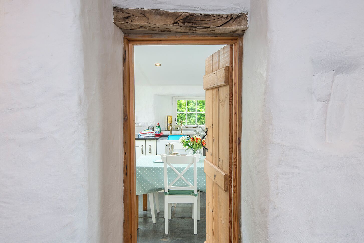 Un estrecho vano de puerta que da a una cocina, con marco de madera, una ventana y una mesa con sillas.
