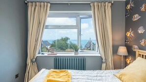 A bedroom with a bed, a window with curtains, a view of the sea, and a lamp. - Robin Hill, Holiday Cottage Tresaith, Sleeps 5, 3 bedrooms,  bathrooms (Cardigan)