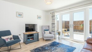A living room with a TV, a grey armchair, a blue sofa, and a large window with a view of a wooden deck. - Sandy-Rise, Holiday Cottage St Davids, Sleeps 4, 2 bedrooms,  bathrooms (Haverfordwest)