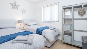 A bedroom with two beds, a nightstand, a lamp, a window with blinds, and a storage unit with baskets. - Sandy-Rise, Holiday Cottage St Davids, Sleeps 4, 2 bedrooms,  bathrooms (Haverfordwest)