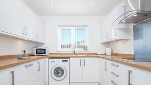 A modern kitchen with white cabinets, a washing machine, a microwave, and a window with blinds. - Sandy-Rise, Holiday Cottage St Davids, Sleeps 4, 2 bedrooms,  bathrooms (Haverfordwest)