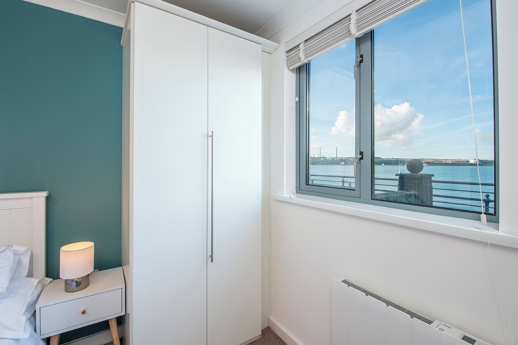 A bedroom with a large window, a bed, a bedside table with a lamp, and a radiator.