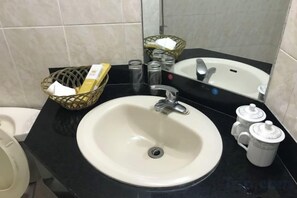 Un baño con un lavamanos blanco, un espejo, una encimera negra, una cesta con artículos de aseo y un pequeño cubo de basura.