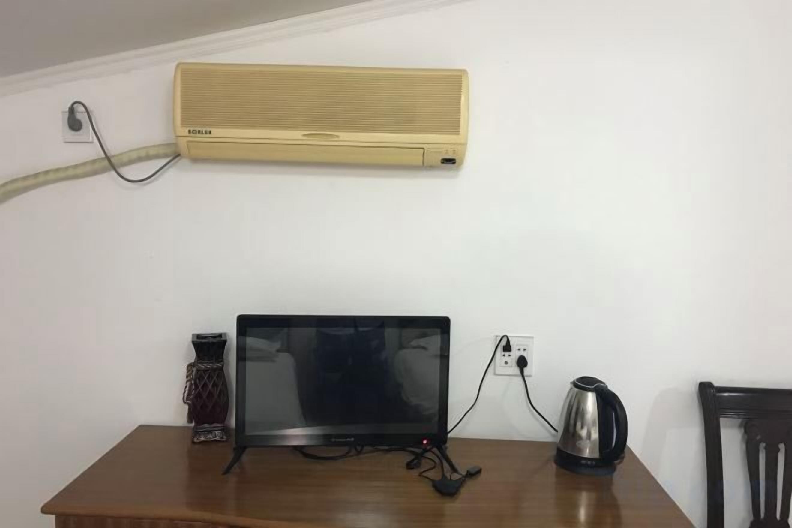 A room with a wooden table, a black monitor, a kettle, and a wall-mounted air conditioner.