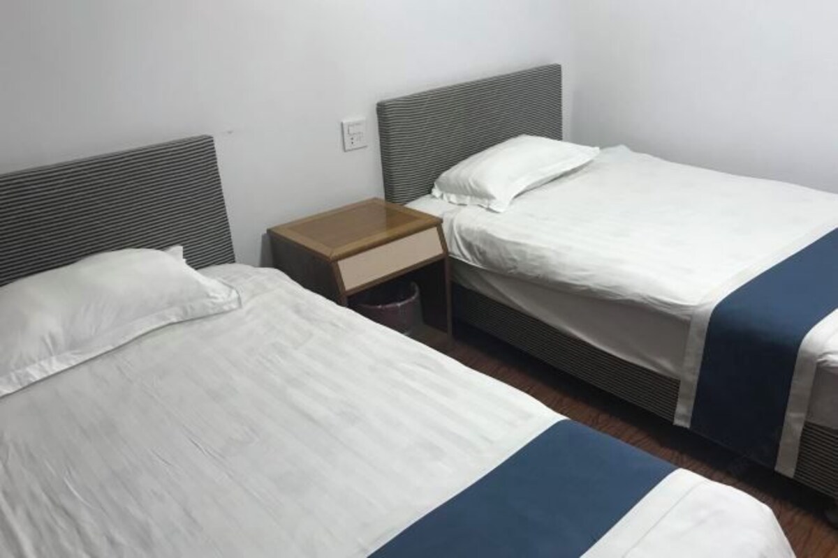 Una habitación de hotel pequeña y con un solo ambiente, con dos camas, cada una con ropa blanca y detalles azules. Hay una mesita de noche de madera con un cesto de basura entre las camas.