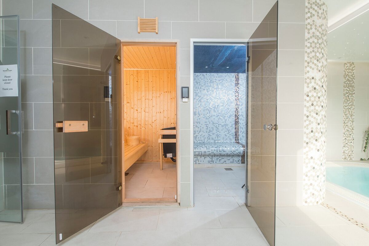 A modern bathroom with a glass shower enclosure, a wooden sauna, and a tiled pool area.