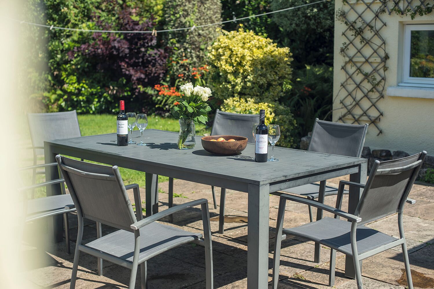 Una mesa para comer al aire libre con seis sillas, un florero con flores, un frutero y dos botellas de vino.