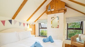 A bedroom with a large bed, wooden beams, a window with a view, and a wicker chair. - Sunnybank, holiday property Manorbier, Sleeps 6, 3 bedrooms,  bathrooms (Manorbier)
