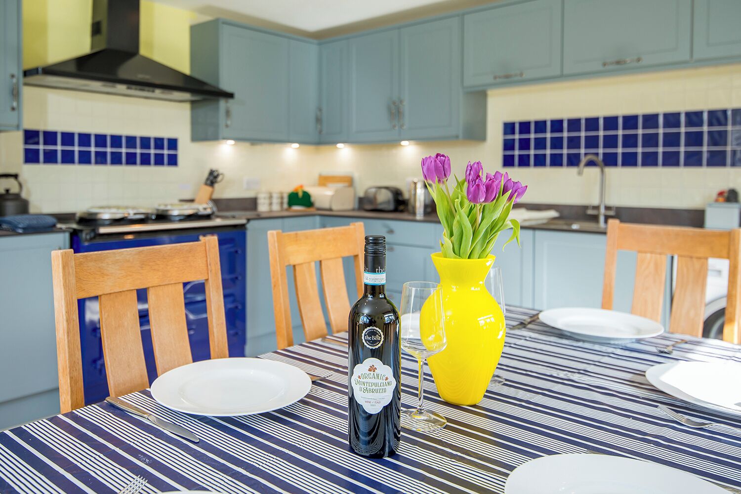 A dining table with a bottle of wine, a yellow vase with flowers, and a striped tablecloth.