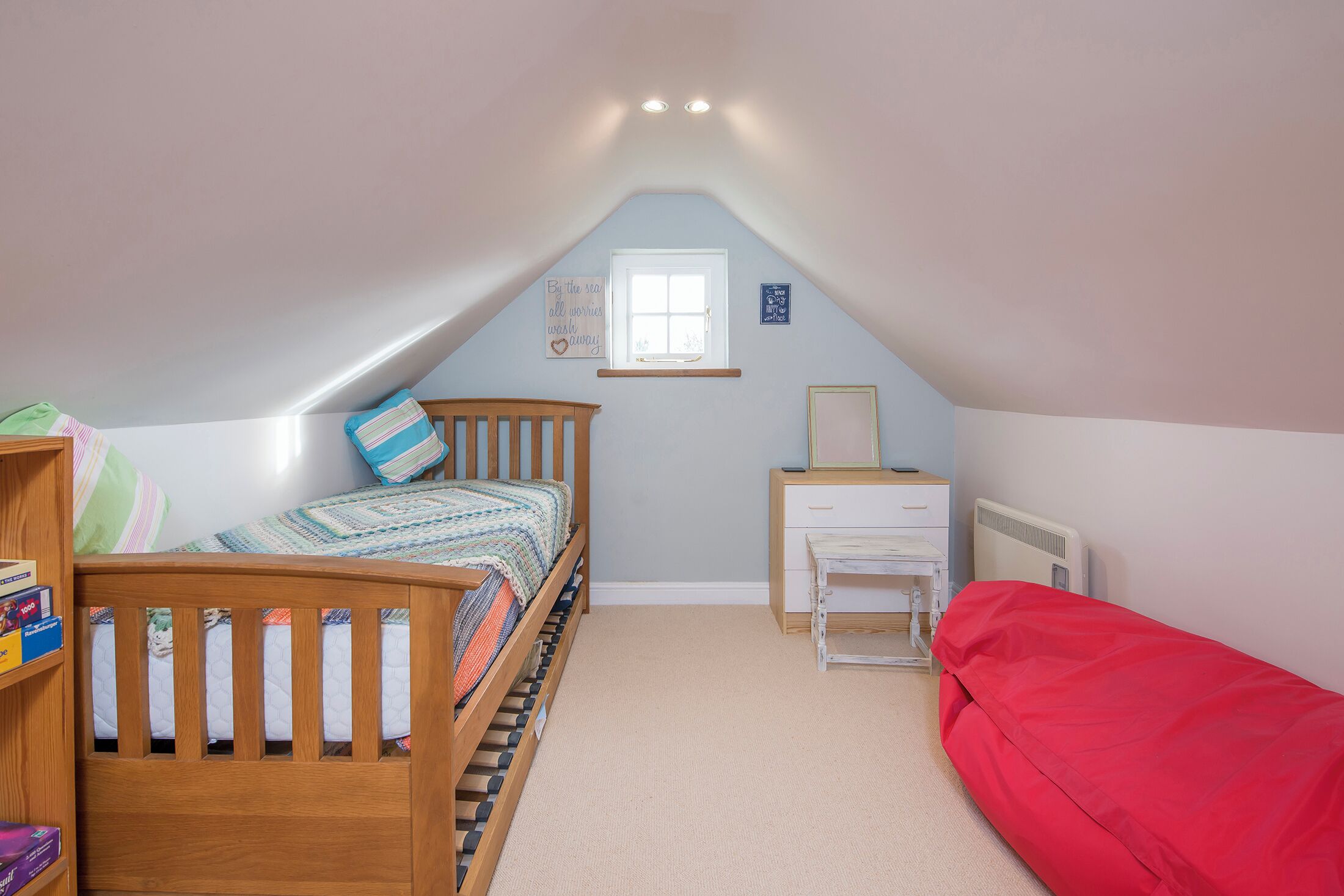 A bedroom with a sloped ceiling, a window, a wooden crib, a red bed, a white dresser, and a radiator.