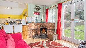 A cozy living room with a fireplace, a red sofa, and a kitchen area with a white counter and a ship's wheel decoration. - Swn y Mor, Holiday Apartment Pwllgwaelod, Sleeps 4, 2 bedrooms,  bathrooms (Dinas)