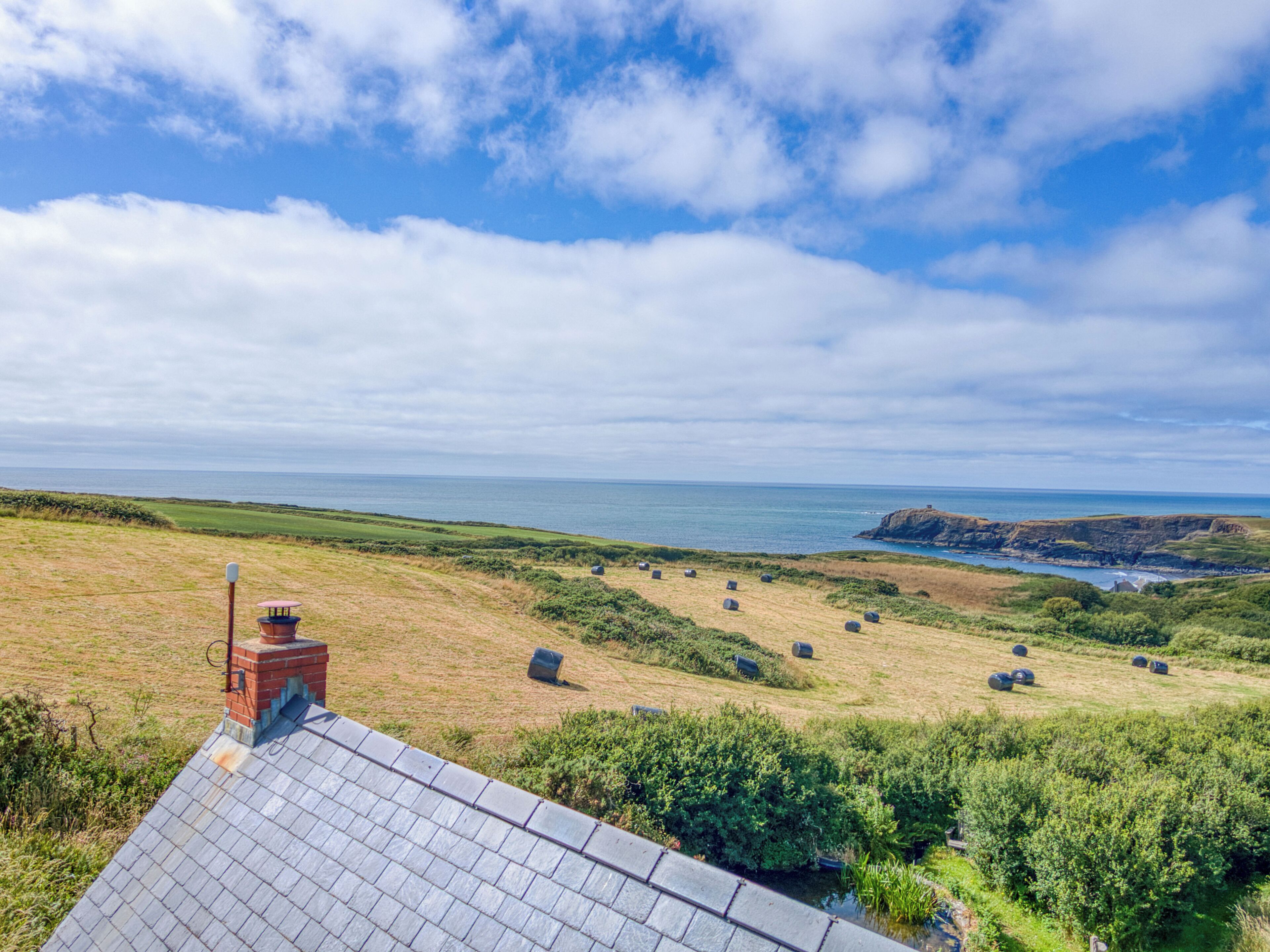 A coastal landscape with a view of the sea, grassy fields, and a building with a sloped roof.