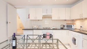A modern kitchen with white cabinets, a built-in oven, microwave, and a washing machine. There is a dining table with chairs and a bottle of wine on it. - The Nook, Holiday Cottage Manorbier, Sleeps 4, 2 bedrooms,  bathrooms (Manorbier)