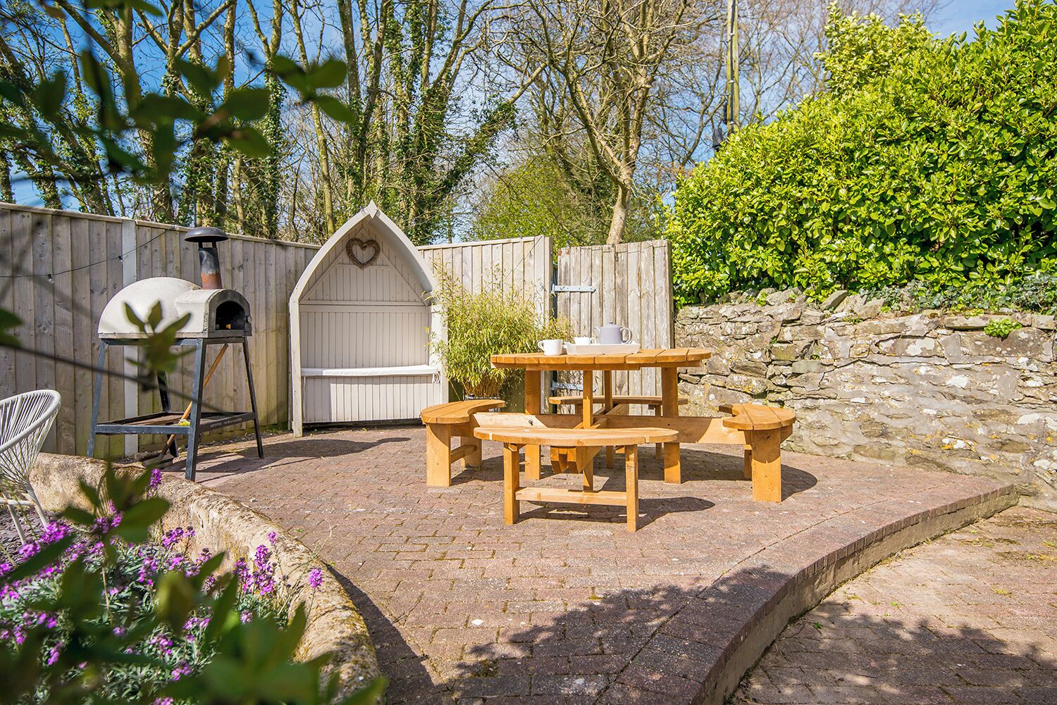 A patio with a wooden table and benches, a white wooden structure with a heart-shaped window, and a stone wall.