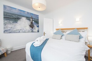 Ein Schlafzimmer mit großem Gemälde, das einen Meerblick zeigt, einem ordentlich bezogenen Bett mit blau-weißer Bettwäsche und einem Holz-Kopfteil.