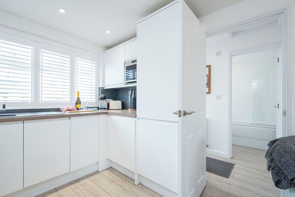 A modern kitchen with white cabinetry, a built-in oven, and a window with blinds. - The Rath Cottage, Holiday Cottage Saundersfoot, Sleeps 2, 1 bedrooms,  bathrooms (SAUNDERSFOOT)