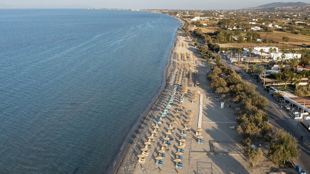 An aerial view of a beach with rows of sun loungers and umbrellas, a road parallel to the beach, and buildings in the background.