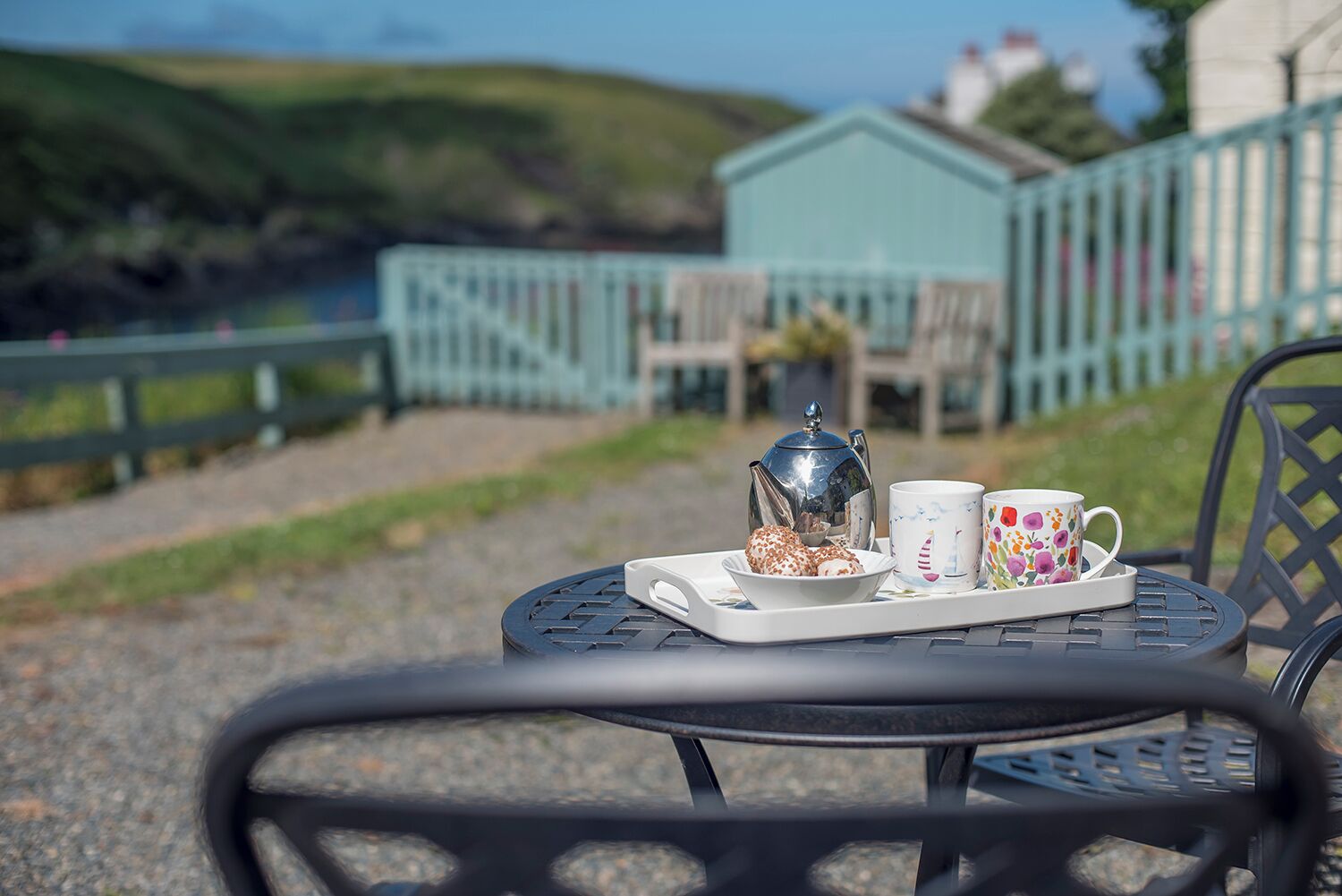 A table with a tray containing a teapot, a mug, and a bowl of pastries, set outdoors with a view of a building and hills in the background.