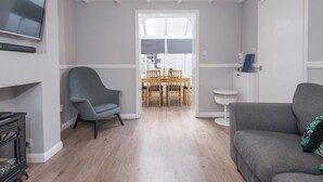 A modern living room with a grey sofa, a grey armchair, a wooden dining table with chairs, and a television mounted on the wall. - The Woolpack, Holiday Cottage Ivy Tower Village, St Florence, Sleeps 6, 3 bedrooms,  bathrooms (Tenby)