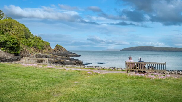 A coastal landscape with a bench overlooking the sea, a grassy area with pink flowers, and rocky terrain. - Tides Reach, Holiday Cottage Cwm yr Eglwys, Sleeps 6, 3 bedrooms,  bathrooms (Newport)