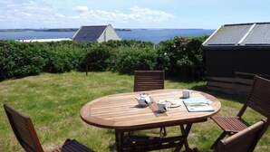 Une table en bois placée à l’extérieur, avec des chaises, une théière, des tasses et un livre, le tout disposé devant un paysage verdoyant, composé de buissons denses et offrant une vue sur la mer et des bâtiments éloignés.
