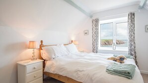 Une chambre avec un lit en bois, du linge de lit blanc, une table de chevet avec une lampe, une fenêtre avec des rideaux et une vue sur la mer.
