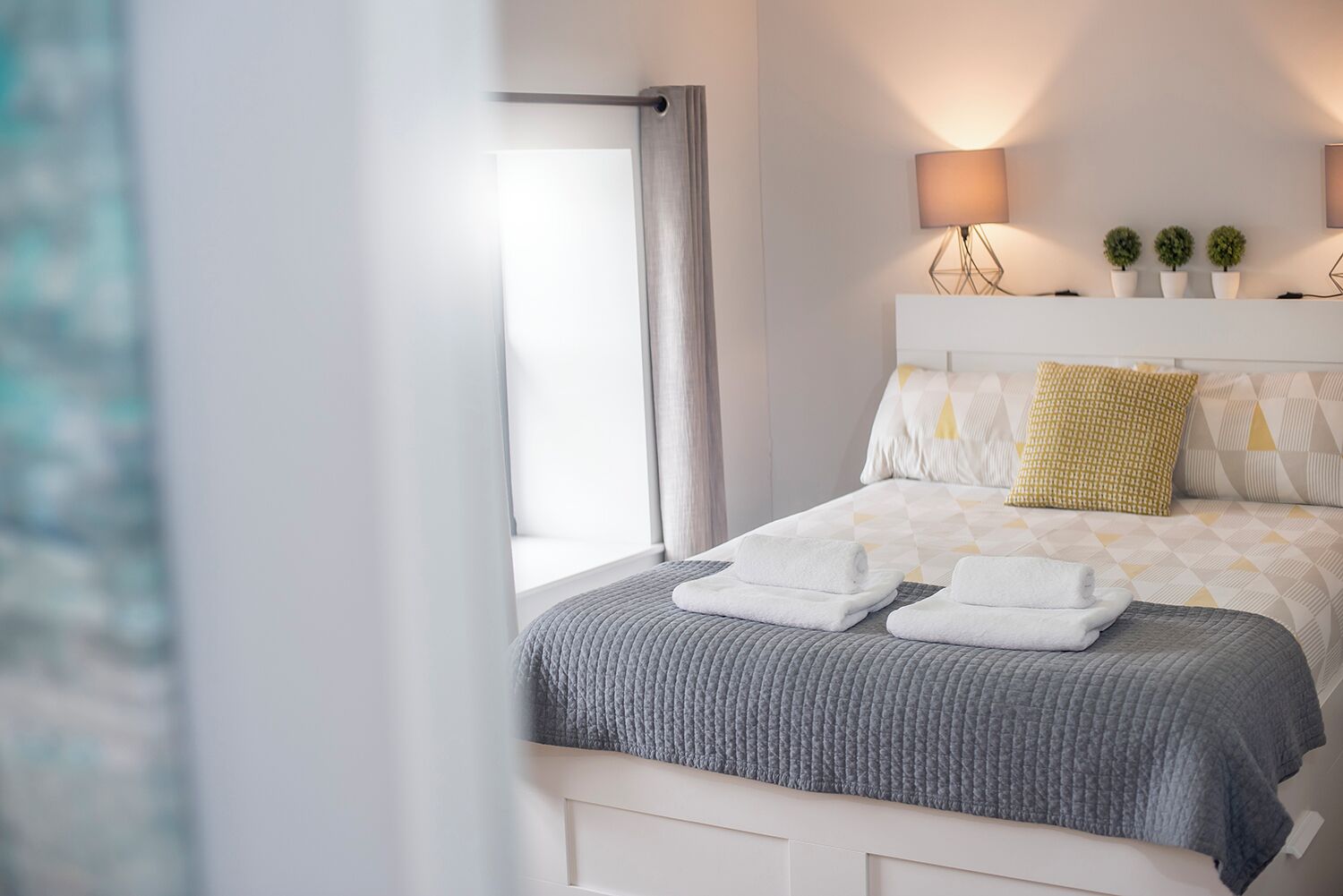 A neatly made bed with a grey blanket and two folded towels, a bedside lamp, and three small potted plants on a white nightstand.