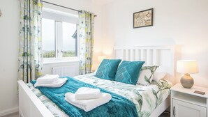 Ein Schlafzimmer mit einem Bett, einem Fenster mit Vorhängen, einem Nachttisch mit Lampe und einem Wandbild.
