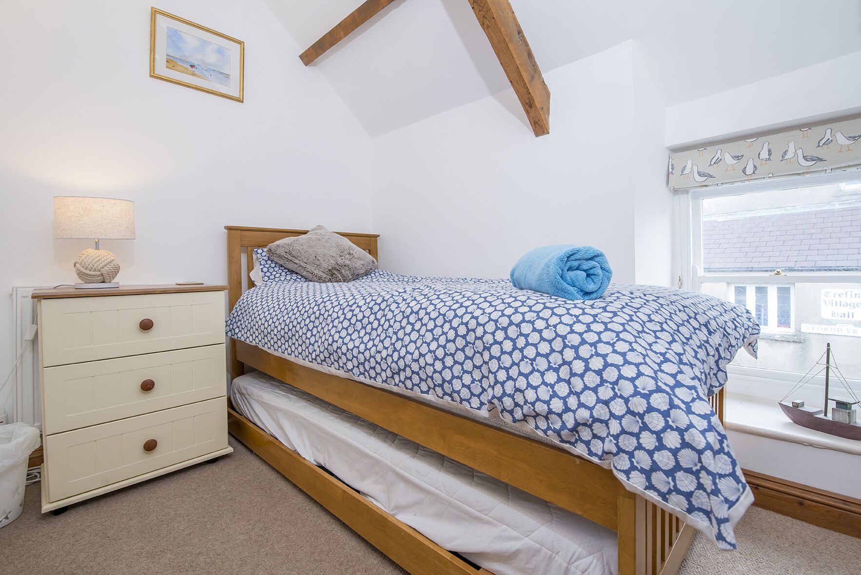 Een slaapkamer met een bed, een dressoir, een lamp, een raam met uitzicht op een dorp, en een ingelijst schilderij aan de muur.
