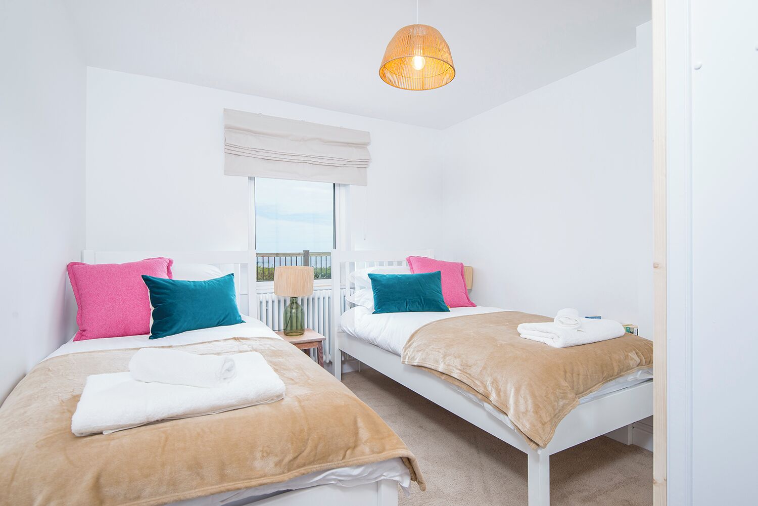 A bedroom with two beds, each with a beige blanket and pillows in pink and teal. There is a window with a view of greenery, a white radiator, and a hanging light fixture.