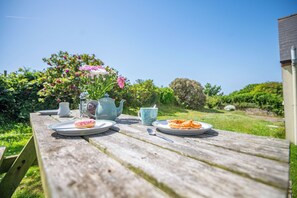 Una mesa de picnic de madera con platos de comida, una tetera y una taza, ubicada al aire libre con un jardín al fondo.