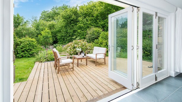 Um deck moderno ao ar livre com piso de madeira, duas cadeiras brancas acolchoadas, uma mesa pequena e uma porta de vidro que dá para um jardim exuberante.