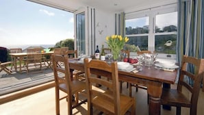 A dining area with a wooden table set for four, large windows with curtains, and a view of the outdoors. - Upper Mead, Holiday Cottage Amroth, Sleeps 6, 3 bedrooms,  bathrooms (NARBERTH)