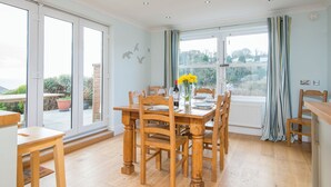 A dining area with a wooden table and chairs, a vase with flowers, and a view through large glass doors. - Upper Mead, Holiday Cottage Amroth, Sleeps 6, 3 bedrooms,  bathrooms (NARBERTH)