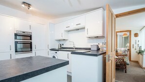 A modern kitchen with white cabinets, a built-in oven, a washing machine, and a dining area with a wicker chair and a table. - Vagwr Eilw Cottage, Holiday Cottage Llanhowell, near Abereiddy, Sleeps 6, 3 bedrooms,  bathrooms (Haverfordwest)