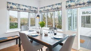 A dining area with a table set for four, large windows with blue floral curtains, a vase with yellow flowers, and a bottle of wine. - Valley View, Holiday Cottage Little Haven, Sleeps 6, 3 bedrooms,  bathrooms (Haverfordwest)