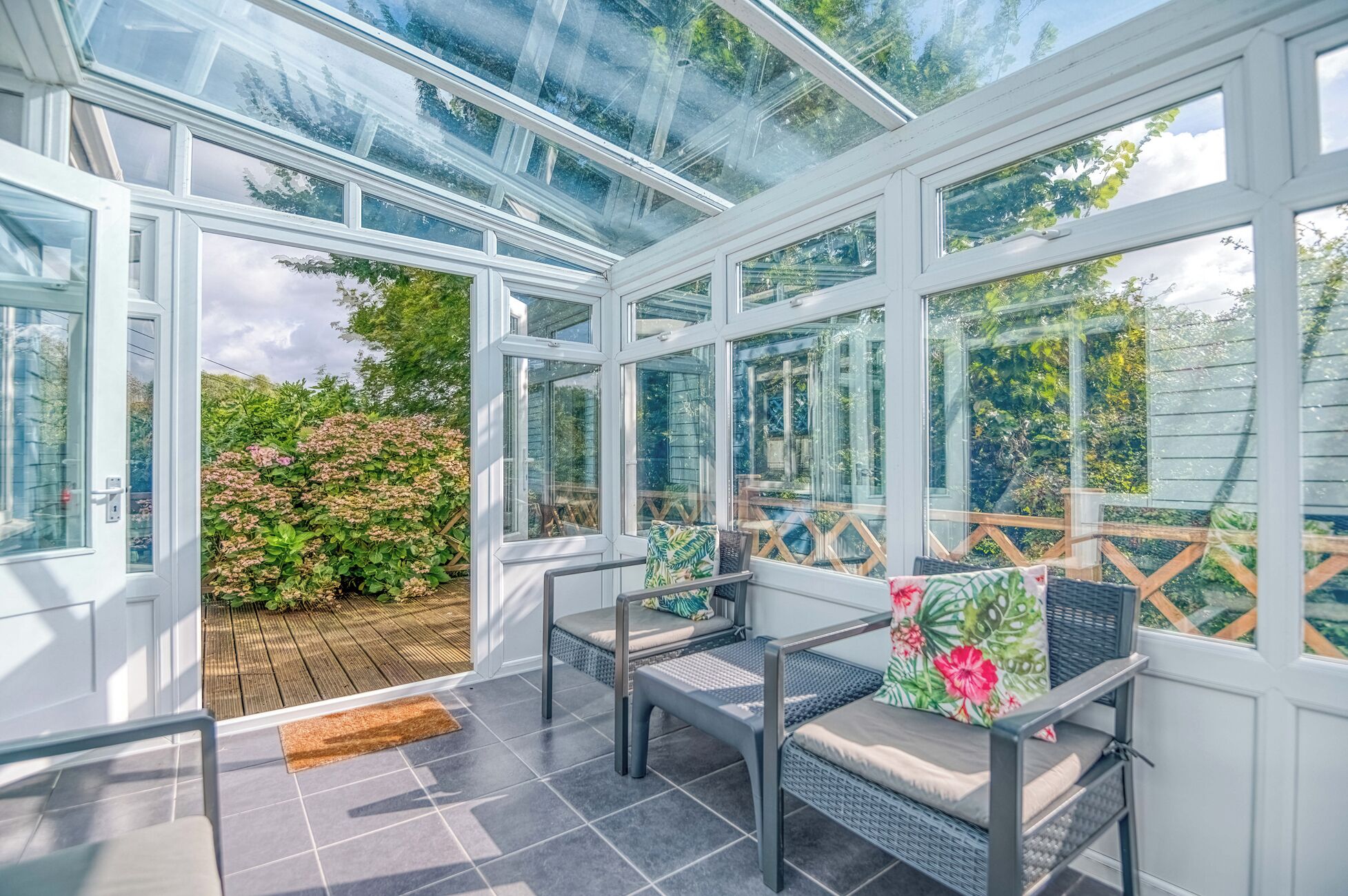 Ein Wintergarten mit Glasdach, zwei Korbstühle mit Kissen und eine Holzterrasse davor.