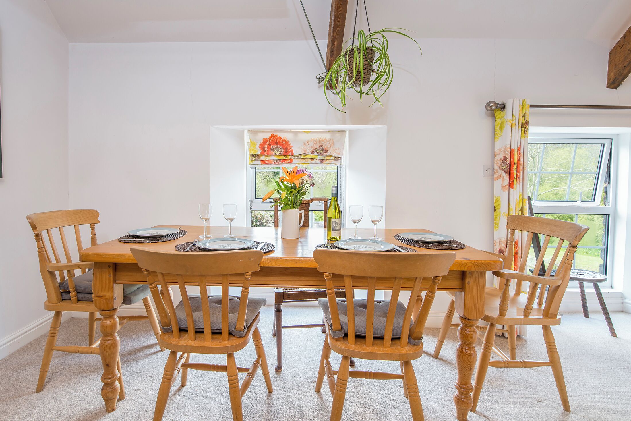 A dining area with a wooden table and chairs, a vase with flowers, and a window with a view of greenery.