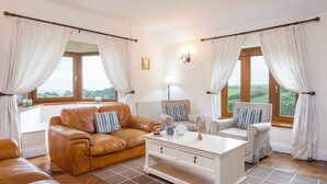 Ein gemütliches Wohnzimmer mit Ledersofa, gestreifte Sessel, einem weißen Couchtisch und Blick ins Freie durch große Fenster.