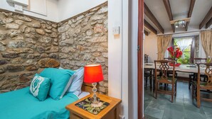 Een slaapkamer met een stenen muur, een bed met blauwgroene beddengoed, een nachtkastje met een lamp, en een zithoek met houten stoelen en een tafel.