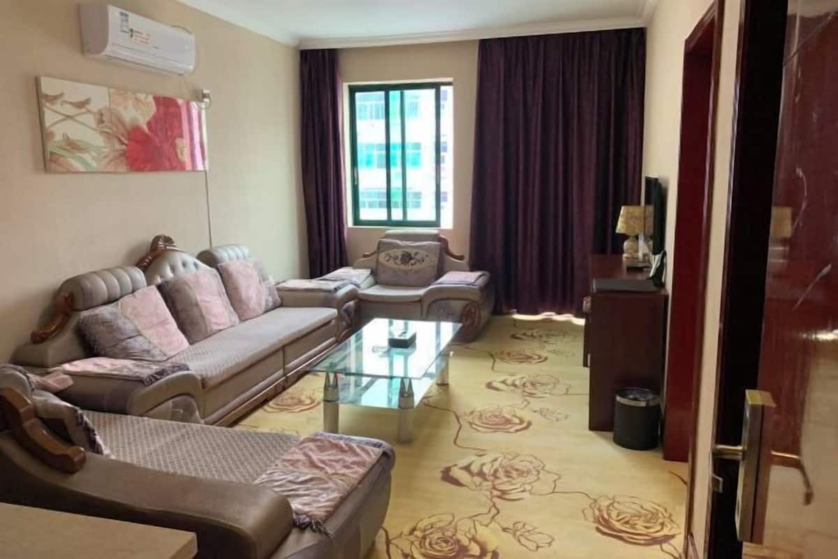 Una sala de estar con sofá, sillones, una mesa de centro de vidrio, una ventana con cortinas y un aire acondicionado de pared.