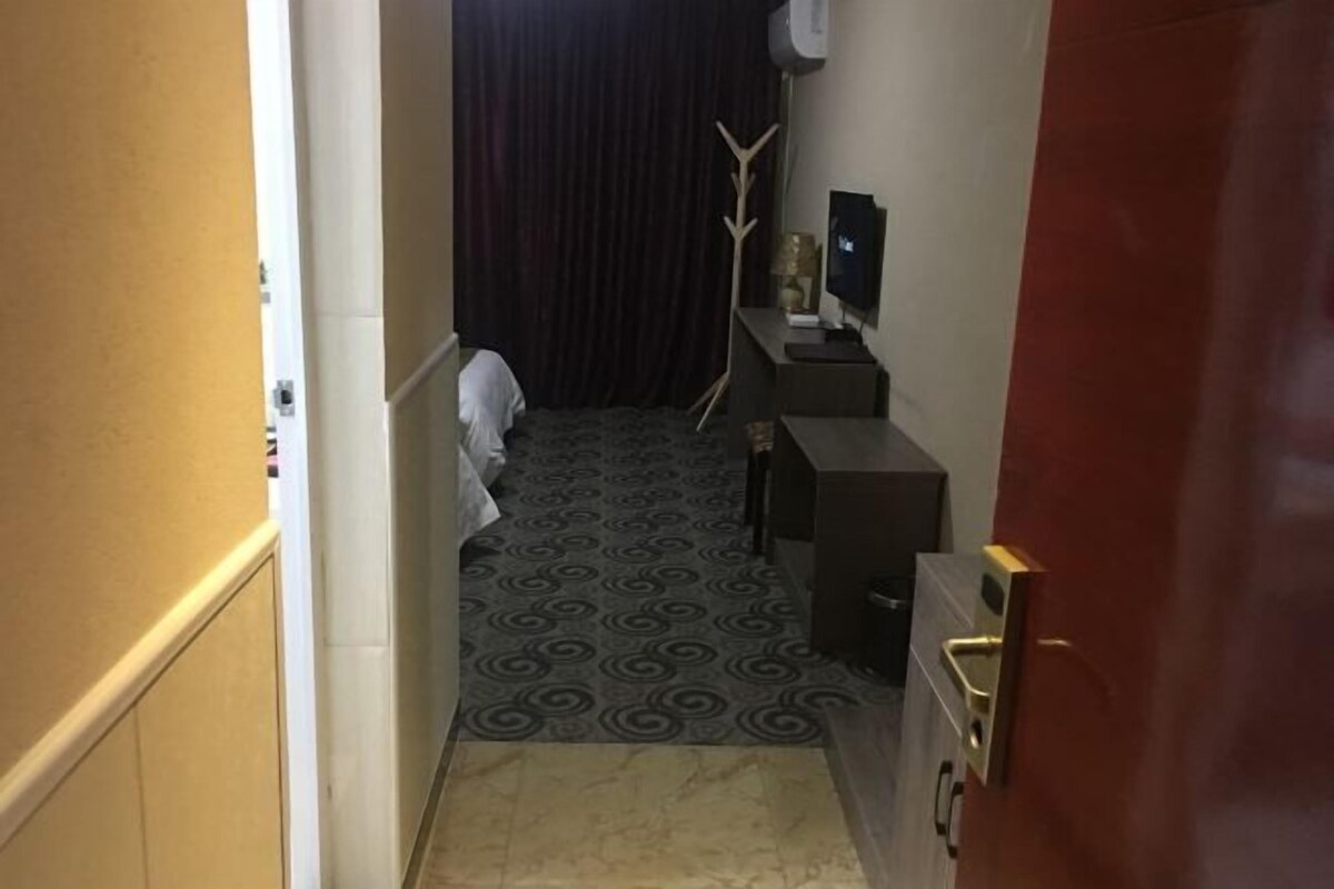 Un pasillo estrecho con alfombra estampada, una cama con cubrecama blanco, un escritorio pequeño con televisor, y una puerta con pomo dorado.