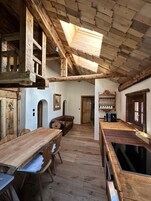 Um interior rústico de madeira com teto alto, vigas de madeira, sala de jantar com mesa e cadeiras, e cozinha com bancada e eletrodomésticos.