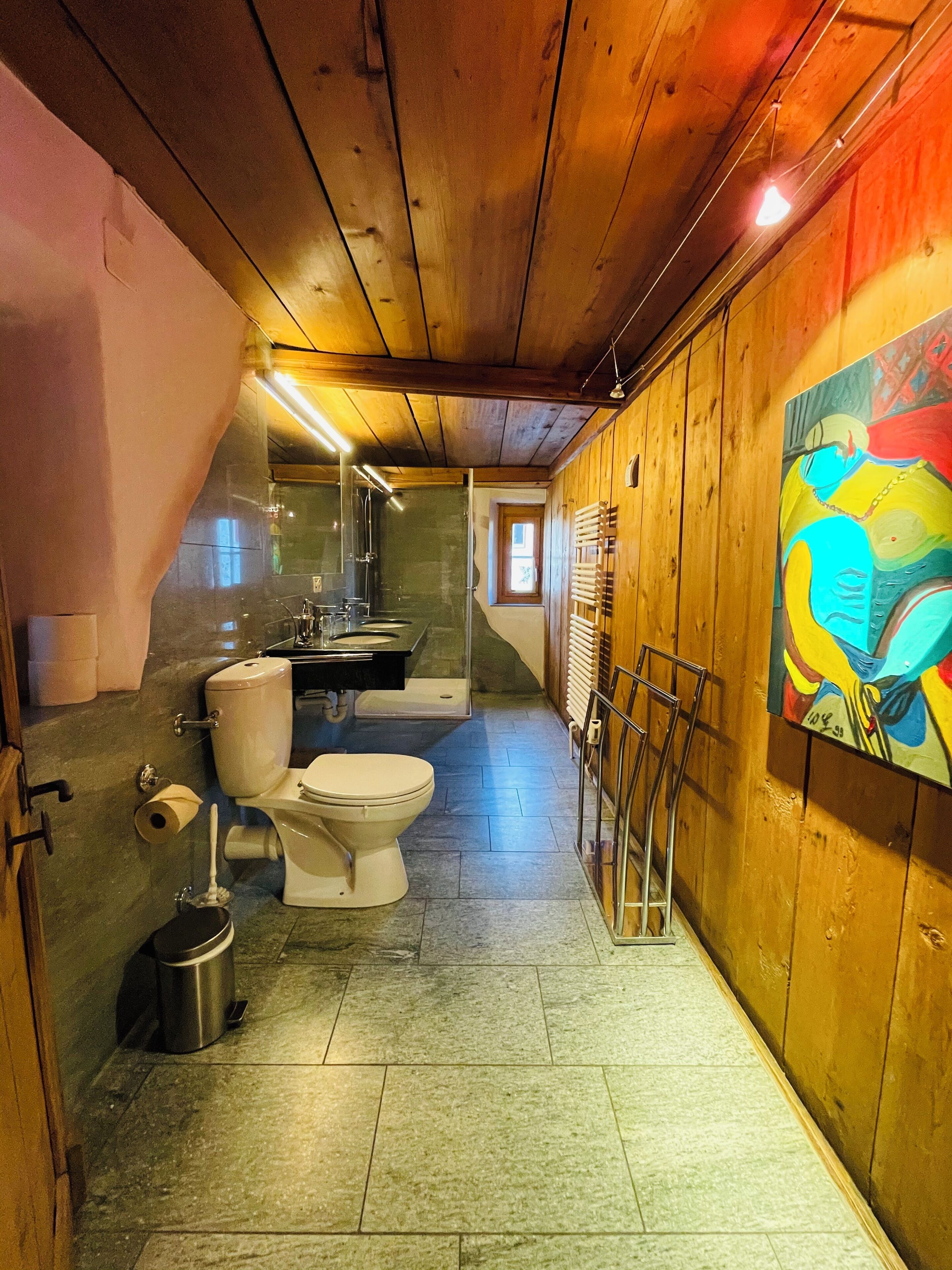 A bathroom with a toilet, bidet, and shower area. There is a painting on the wall and a window in the background.